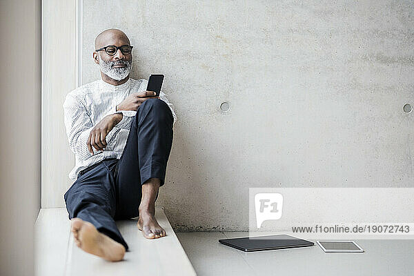 Barefoot mature businessman sitting on window sill looking at smartphone