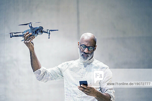 Portrait of bald mature man holding quadcopter while looking at cell phone