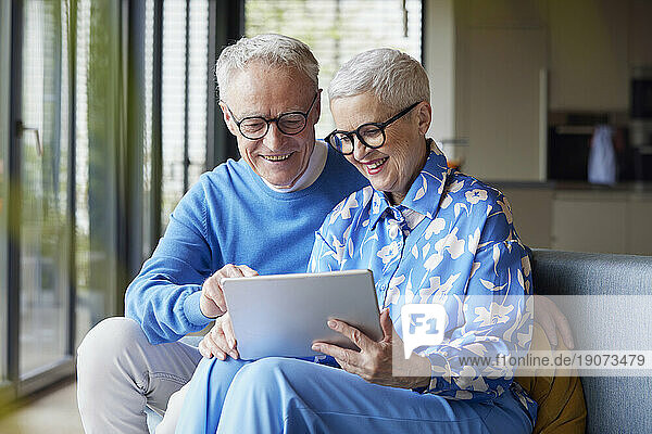 Happy senior couple sitting on couch at home using tablet PC