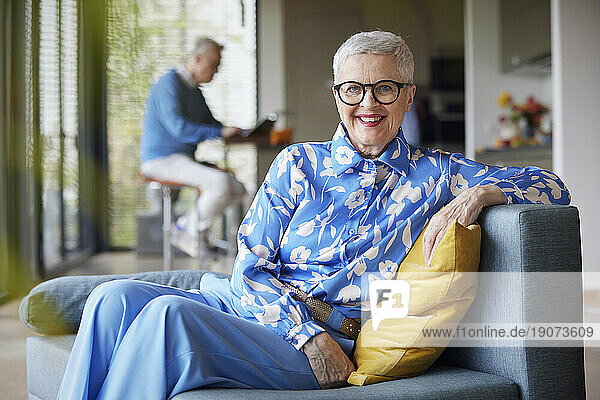 Smiling senior woman sitting on couch at home with man in background
