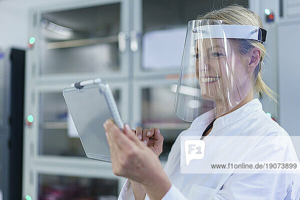 Smiling mature female scientist wearing protective face shield while using digital tablet at laboratory