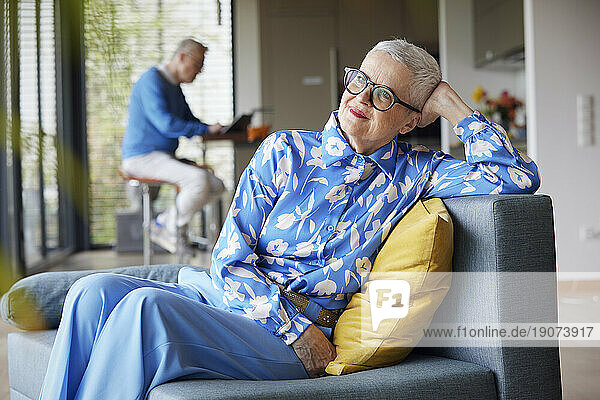 Relaxed senior woman sitting on couch at home with man in background