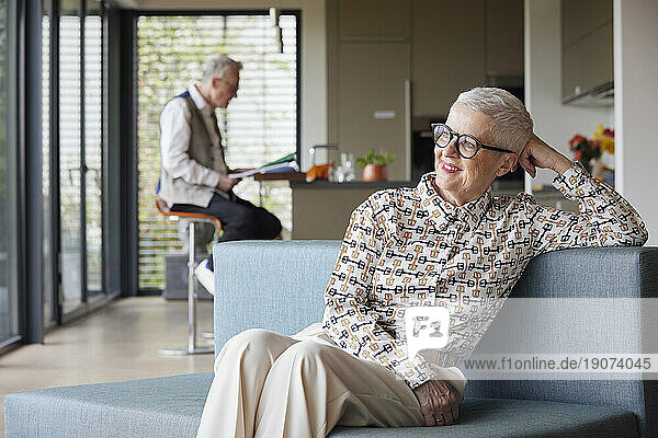 Smiling senior woman sitting on couch at home with man in background