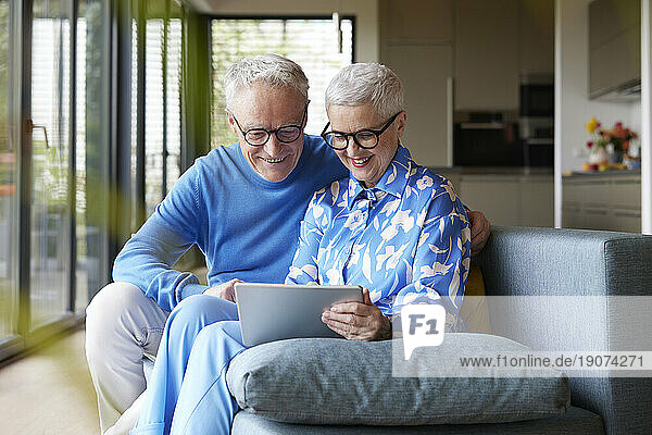 Happy senior couple sitting on couch at home using tablet PC