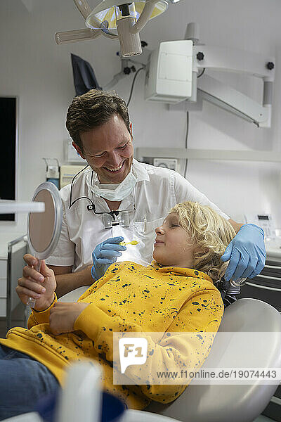 Blond boy looking in hand mirror at dental clinic