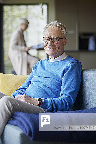 Portrait of smiling senior man sitting on couch at home with woman in background