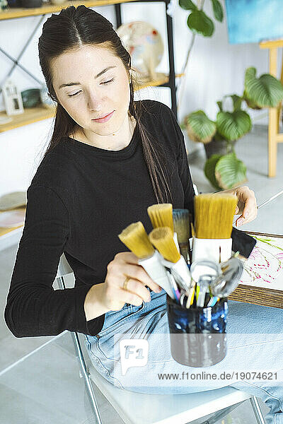 Contemplative artist choosing paintbrush sitting on chair in workshop