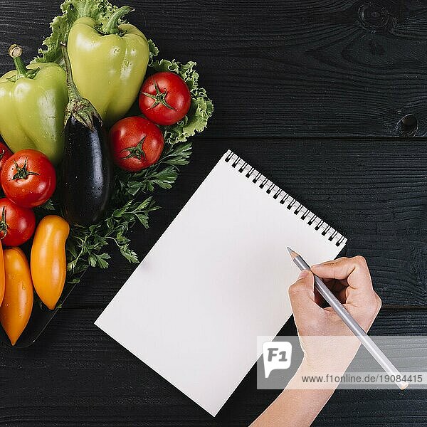Menschliche Hand schreiben Spirale Notizblock mit frischem Gemüse schwarzem Holzhintergrund