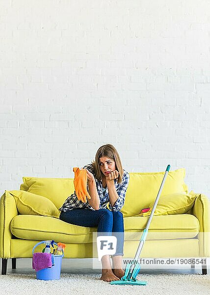 Traurige junge Frau sitzt auf einem gelben Sofa und schaut auf orangefarbene Gummihandschuhe