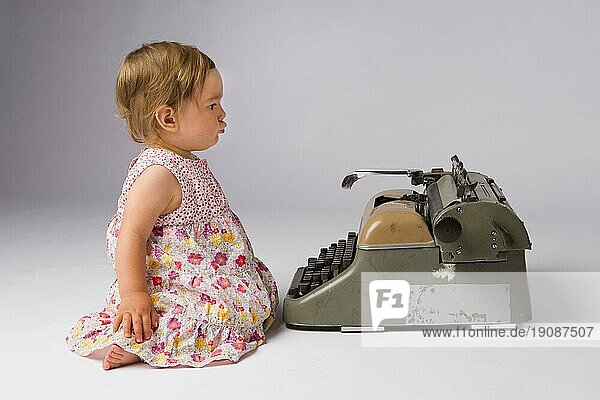 Nachdenklicher Ausdruck eines niedlichen einjährigen Mädchens  das vor einer Schreibmaschine sitzt und überlegt  was es damit anfangen soll