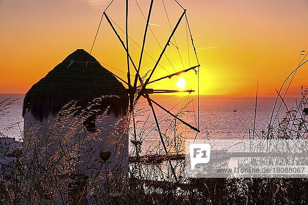 Berühmte traditionelle griechische Windmühle auf der Spitze des Hügels Mykonos  Kykladen  Griechenland bei Sonnenuntergang. Schöne Sonnenuntergang Himmel  Sonne berühren Meer Horizont  hohes Gras im Vordergrund  buntes Bild  selektiver Fokus  unterbelichtet
