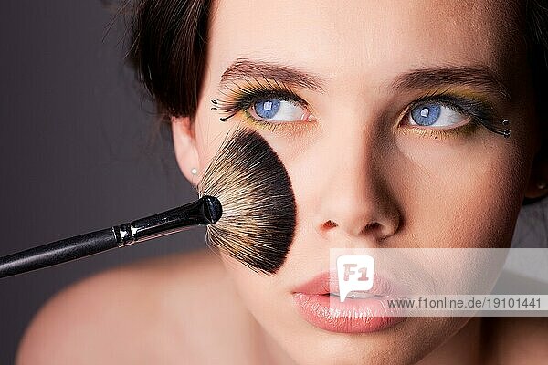Beauty Shooting einer jungen Frau mit langen Wimpern  blaün Augen und Schminkpinsel  der ihr Gesicht berührt. Geringe Tiefenschärfe