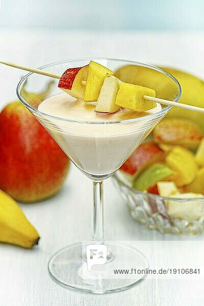 Ein Glas mit Mango Birne Shake. Diagonal über dem Glas liegen mehrere Fruchtstücke  die auf einen Stab aufgespießt sind. Der Unter- Hintergrund ist bewußt unscharf gehalten