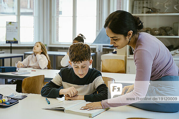 Eine Lehrerin hilft einem Schüler beim Lesen in einem Buch  während er auf dem Schreibtisch im Klassenzimmer sitzt
