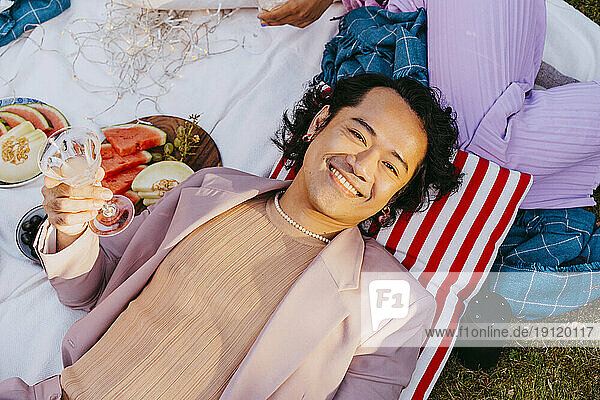 High angle portrait of smiling gay man holding wineglass while lying down in back yard