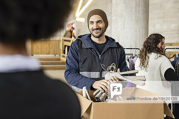 Lächelnder männlicher Arbeiter beim Auspacken von Kleiderbügeln aus einem Karton im Recyclingzentrum