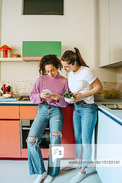 Junge Frau  die ihr Smartphone mit einer Freundin teilt  während sie sich zu Hause auf die Küchentheke stützt