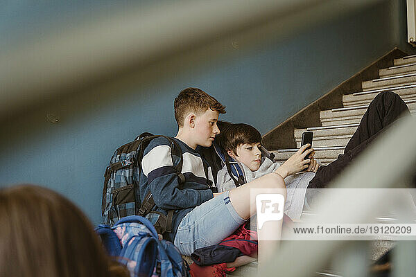 Männliche Freunde teilen sich ein Smartphone  während sie auf der Treppe eines Schulgebäudes sitzen