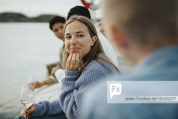 Lächelnde junge Frau mit Hand am Kinn  die mit Freunden auf einem Steg am See sitzt