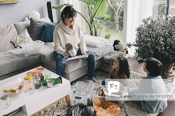 Lächelnde Frau mit Sohn und Tochter  die im Wohnzimmer sitzend den Müll trennen