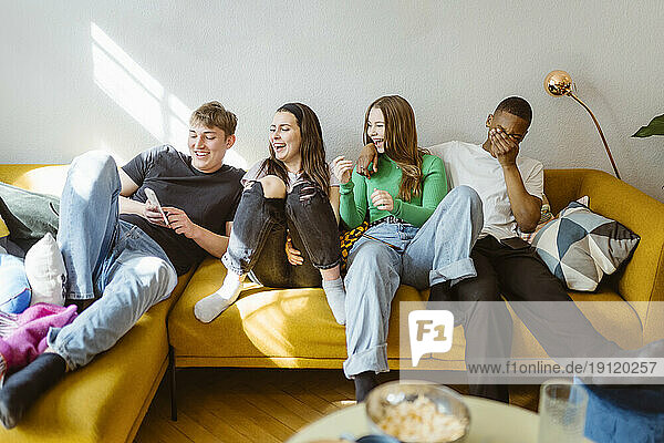 Fröhliche junge gemischtrassige Freunde benutzen ihr Smartphone  während sie zu Hause auf dem Sofa sitzen