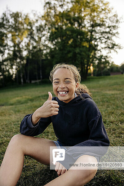 Portrait of happy girl showing thumbs up while sitting on grass in playground