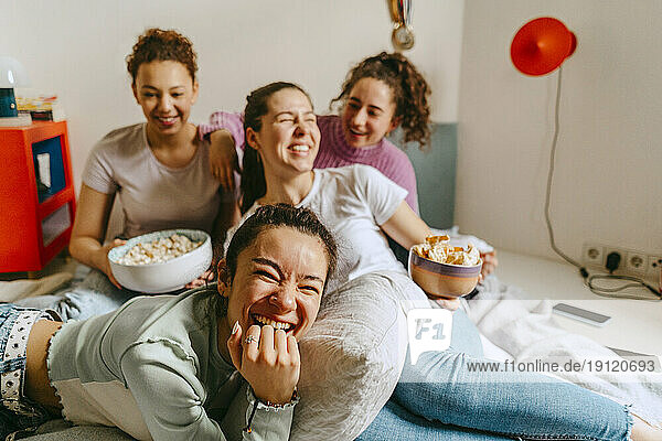 Cheerful female friends watching TV while enjoying snacks at home