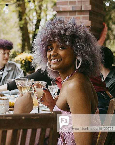Seitenansicht Porträt der lächelnden Transfrau mit lockigem Haar sitzt mit Freunden während der Party im Hinterhof