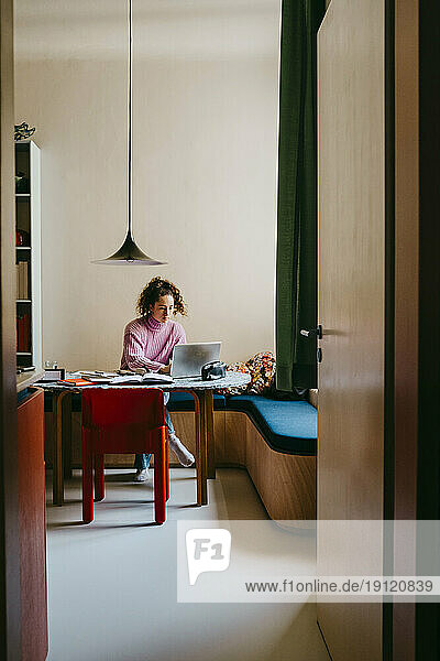 Gesamtansicht einer jungen Frau  die einen Laptop benutzt  während sie am Tisch Hausaufgaben macht