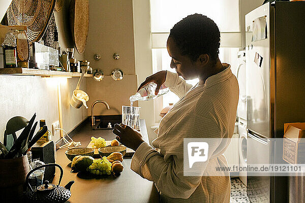 Side view of woman wearing bathrobe pouring water in glass at kitchen counter