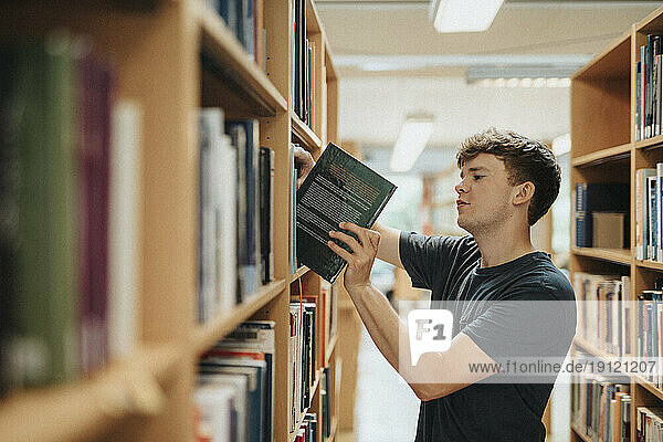 Seitenansicht eines männlichen Studenten  der ein Buch aus einem Bücherregal in der Bibliothek einer Universität entnimmt