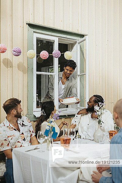Lächelnder Mann  der Freunden am Tisch sitzend während einer Dinnerparty Kuchen schenkt