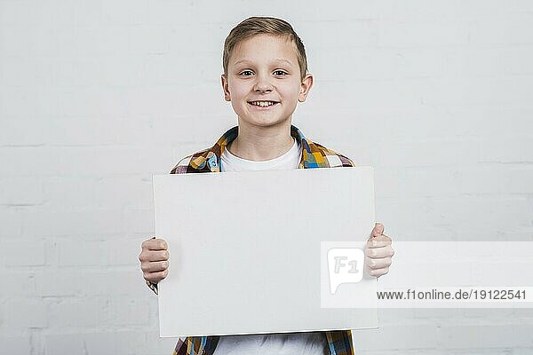 Porträt glücklicher Junge  der vor einer weißen Wand steht und ein weißes leeres Plakat zeigt