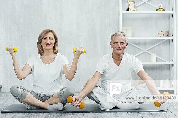 Lächelndes Paar sitzt auf einer Yogamatte und übt mit Hanteln
