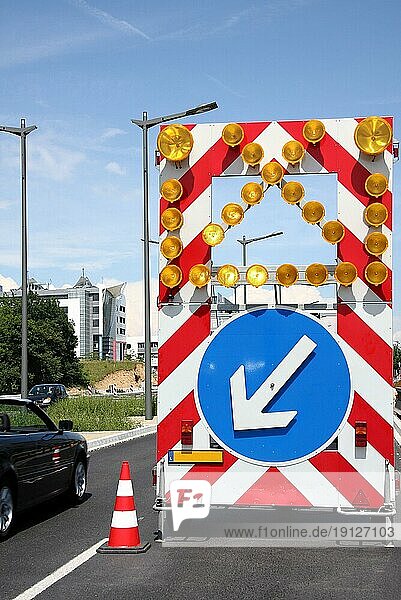 Verkehrstafel mit Warnleuchten und blauem Verkehrsschild mit Pfeil-nach-links  Hinweis zur Benutzung des anderen Fahrstreifens  zwei Autofahrer auf zwei verschiedenen Fahrbahnen  Hintergrund Gebäude und Himmel