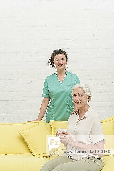 Lächelnde ältere Frau sitzt auf dem Sofa und hält eine Kaffeetasse vor der Krankenschwester