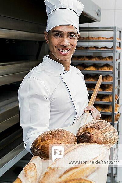 Porträt lächelnder junger männlicher Bäcker mit frisch gebackenem Brot und Holzschaufel