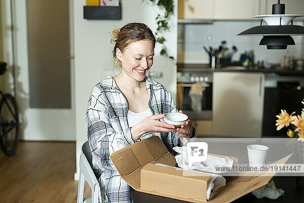 Smiling young woman unpacking ceramic bowls from cardboard box at home