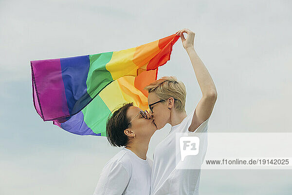 Loving lesbians with rainbow flag kissing each other under sky
