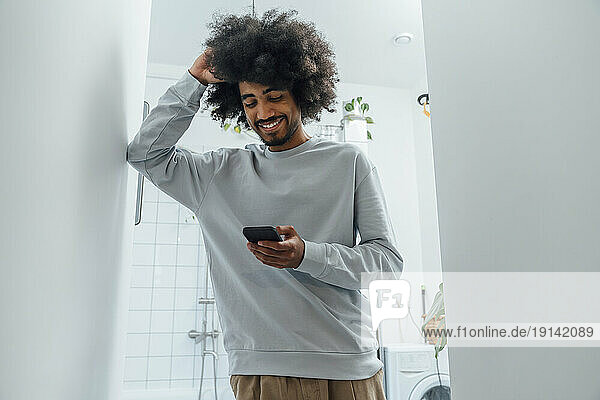 Smiling man with hand in hair using smart phone in bathroom at home
