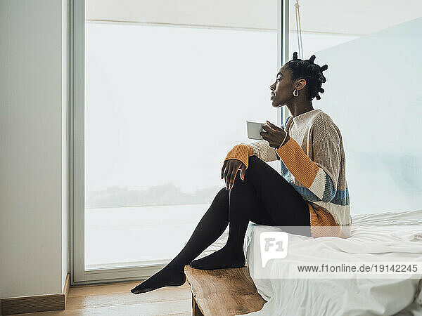 Young woman having coffee sitting on bed near window