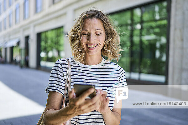 Happy woman with blond hair using smart phone near building