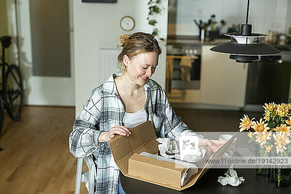 Smiling young woman unpacking ceramic bowls from cardboard box on table