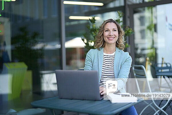 Smiling freelancer sitting with laptop at sidewalk cafe