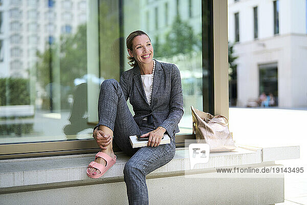 Smiling businesswoman with book sitting on bench near glass wall