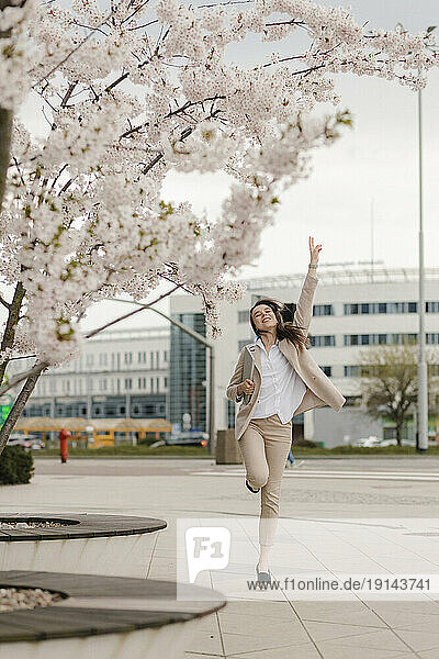 Cheerful young businesswoman jumping by cherry blossom flower tree