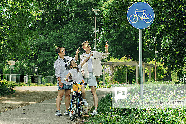 Lesbian couple with daughter pointing at bicycle lane sign in park