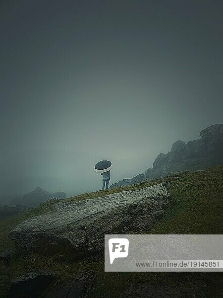 Rückansicht eines einsamen Mannes mit Regenschirm steht auf einem felsigen Hügel  der von Dunst bedeckt ist. Stimmungsvolle und emotionale Szene mit einem einsamen Fremden Silhouette unter dem regen
