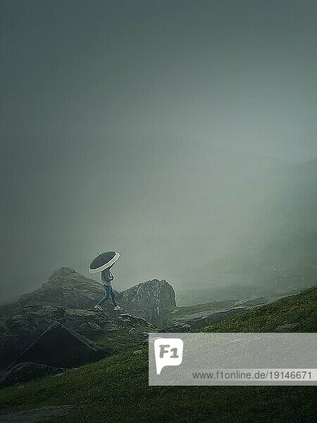 Mann mit Regenschirm  der bei düsterem Wetter und dichtem Nebel einen Berg hinaufsteigt. Stimmungsvolle Wanderszene mit einem Wanderer  der auf den Klippen des nebligen Tals wandert
