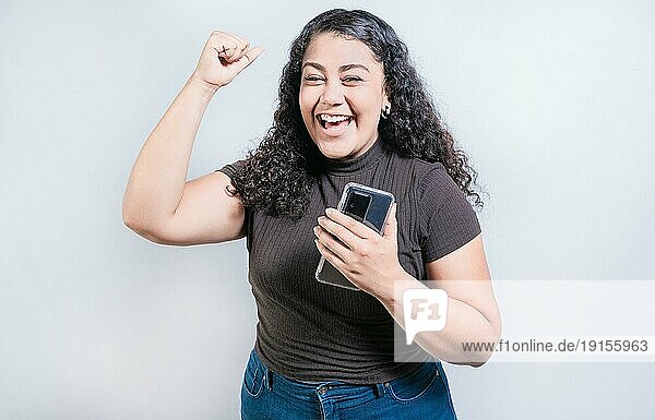 Winning glückliche junge Frau hält Handy. Happy latin girl celebrating with phone isolated. Glückliche Menschen halten Smartphone und feiern